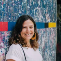 Carol é uma mulher branca de cabelos castanhos de tamanho médio, ela usa uma argola grande e sorri