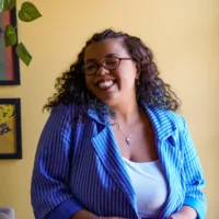 Gabriela é uma mulher negra, de cabelos cacheados longo, que usa óculos e um blazer azul listrado