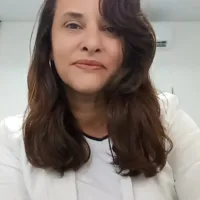 Taty é uma mulher branca de cabelos castanhos de tamanho médio. Ela usa uma blusa e um blazer brancos.
