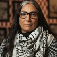 Soraya é uma mulher de ascendência palestina, de cabelos longos com raízes grisalhas, que usa óculos e um lenço palestino no pescoço