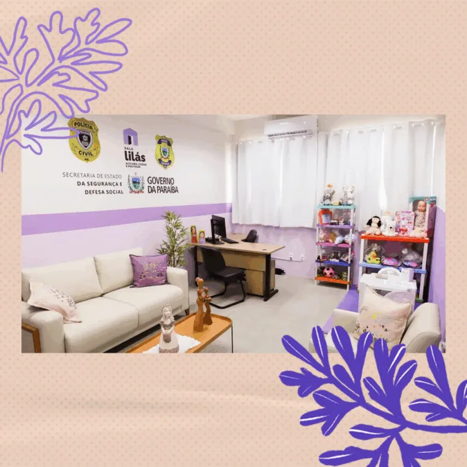A imagem mostra uma sala organizada e acolhedora, com paredes brancas e uma faixa horizontal lilás. Na parede à esquerda, estão os logotipos da Polícia Civil, da “Sala Lilás”, da Secretaria de Estado da Segurança e Defesa Social e do Governo da Paraíba.

No canto esquerdo, há um sofá branco com almofadas lilases e uma mesa de centro de madeira com duas esculturas decorativas. À frente, uma mesa de escritório de madeira clara e estrutura preta, com um computador e duas cadeiras, uma de cada lado.

No canto direito da sala, há duas estantes coloridas cheias de brinquedos, bonecas e pelúcias. As janelas ao fundo estão cobertas por cortinas brancas, e um ar-condicionado fica acima delas.
