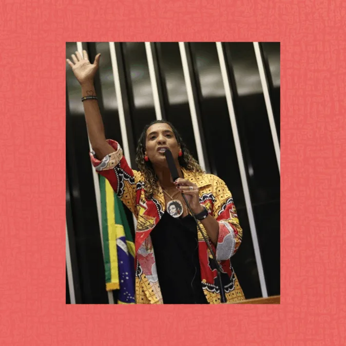 Mulher negra fala ao microfone em plenário, braço esquerdo erguido. Usa casaco estampado colorido sobre blusa preta e colar com pingente redondo. Ao fundo, faixas verticais escuras e a bandeira do Brasil parcialmente visível. Enquadramento de baixo para cima, gesto firme, clima de pronunciamento.