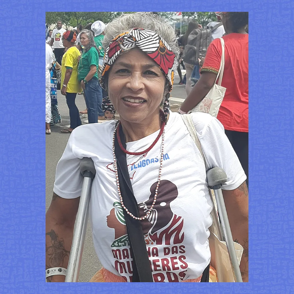 Mulher sorrindo em ambiente de marcha, vestindo camiseta branca estampada, colar de contas e turbante colorido. Ela apoia-se em duas muletas metálicas e carrega uma bolsa no ombro. Ao fundo, outras pessoas caminham pela rua