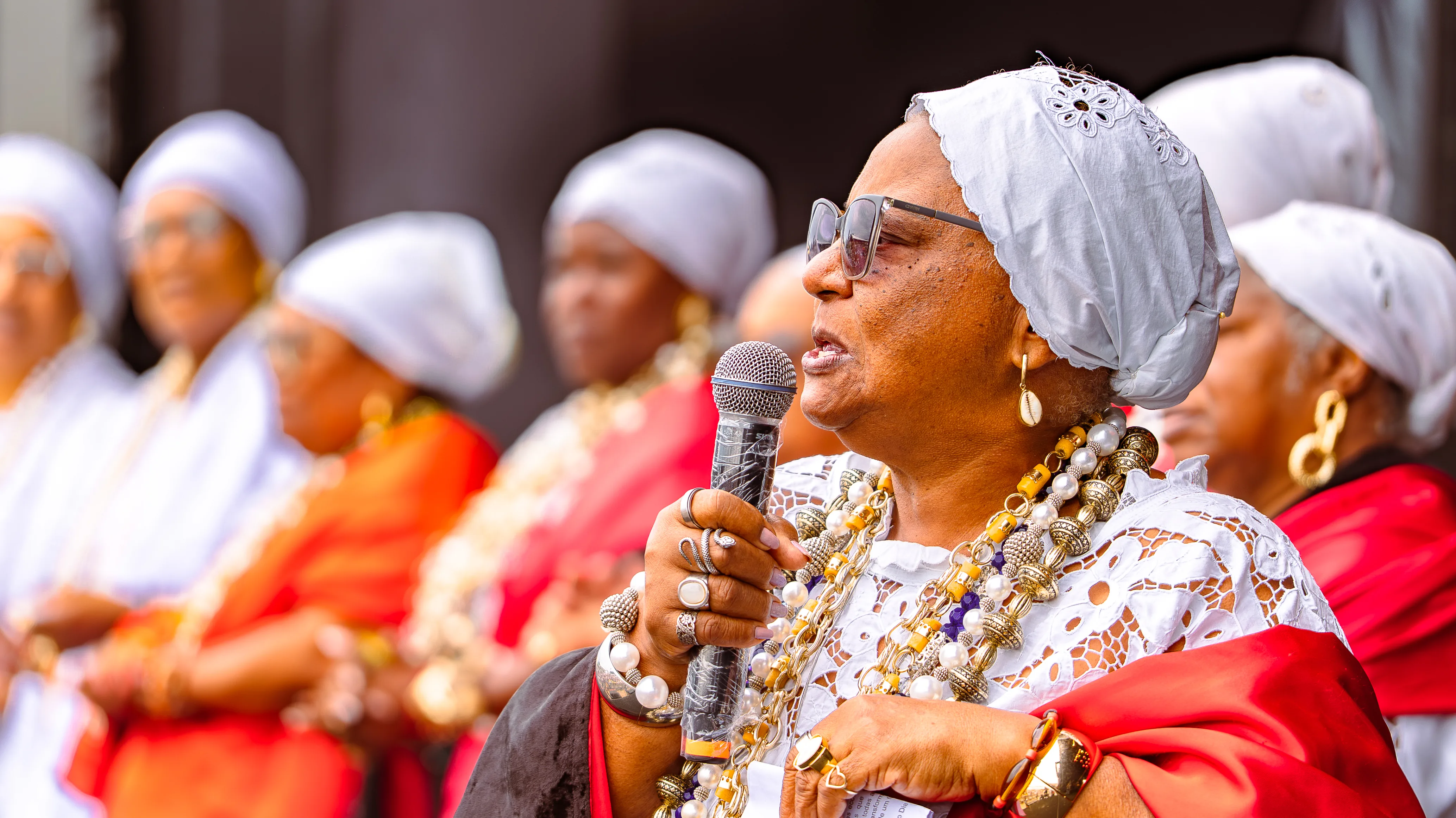 Mulher com turbante branco, óculos e colares longos fala ao microfone enquanto outras mulheres alinhadas ao fundo, também de turbante e roupas brancas e vermelhas, observam.