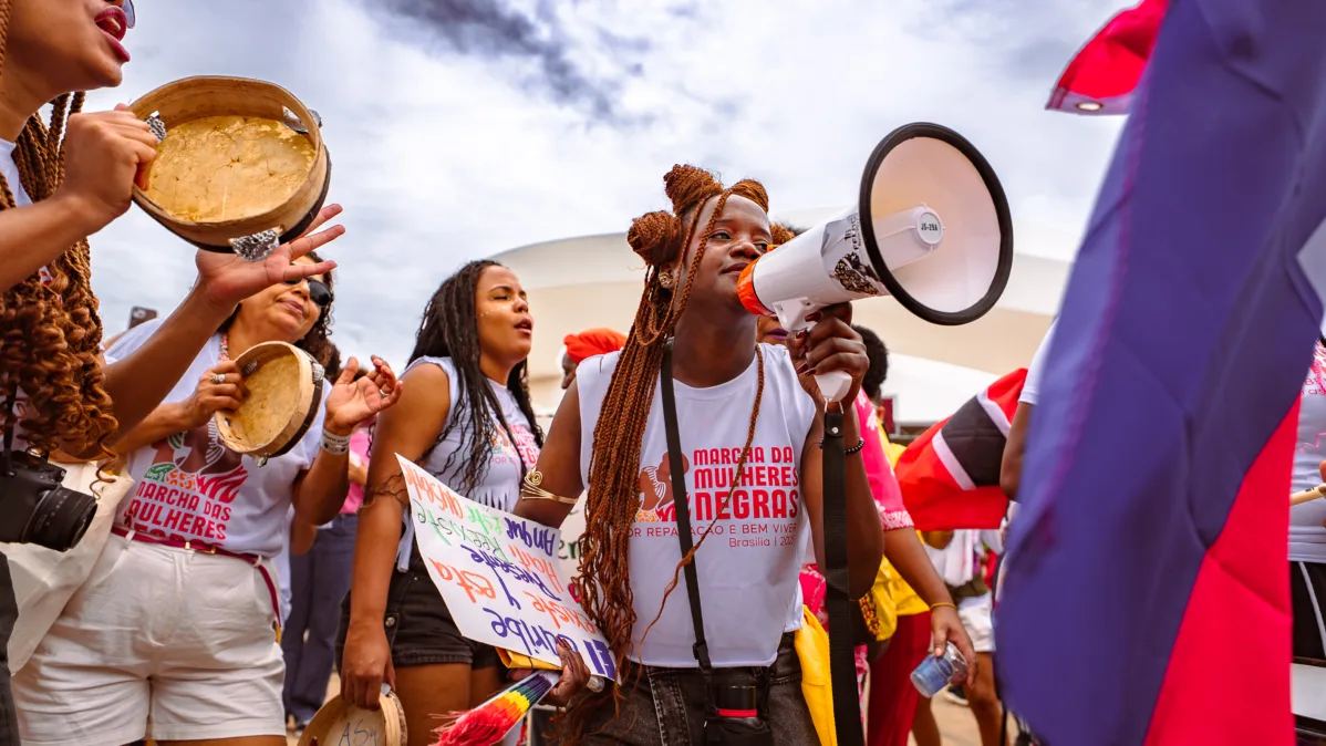 Mulher com tranças longas segura megafone enquanto outras pessoas ao redor tocam pandeiros e carregam cartazes. Todos vestem camisetas da marcha.