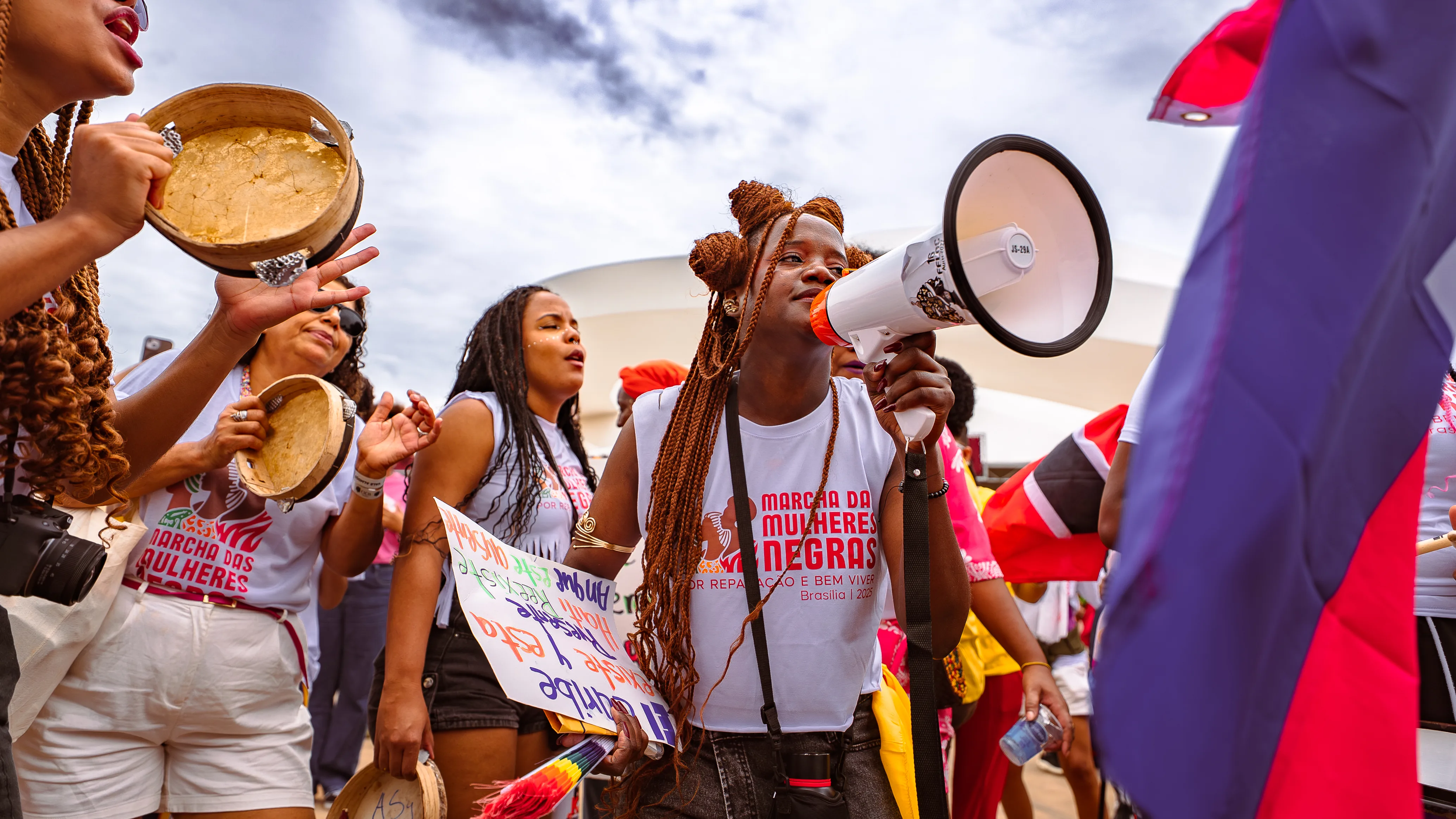 Mulher com tranças longas segura megafone enquanto outras pessoas ao redor tocam pandeiros e carregam cartazes. Todos vestem camisetas da marcha.