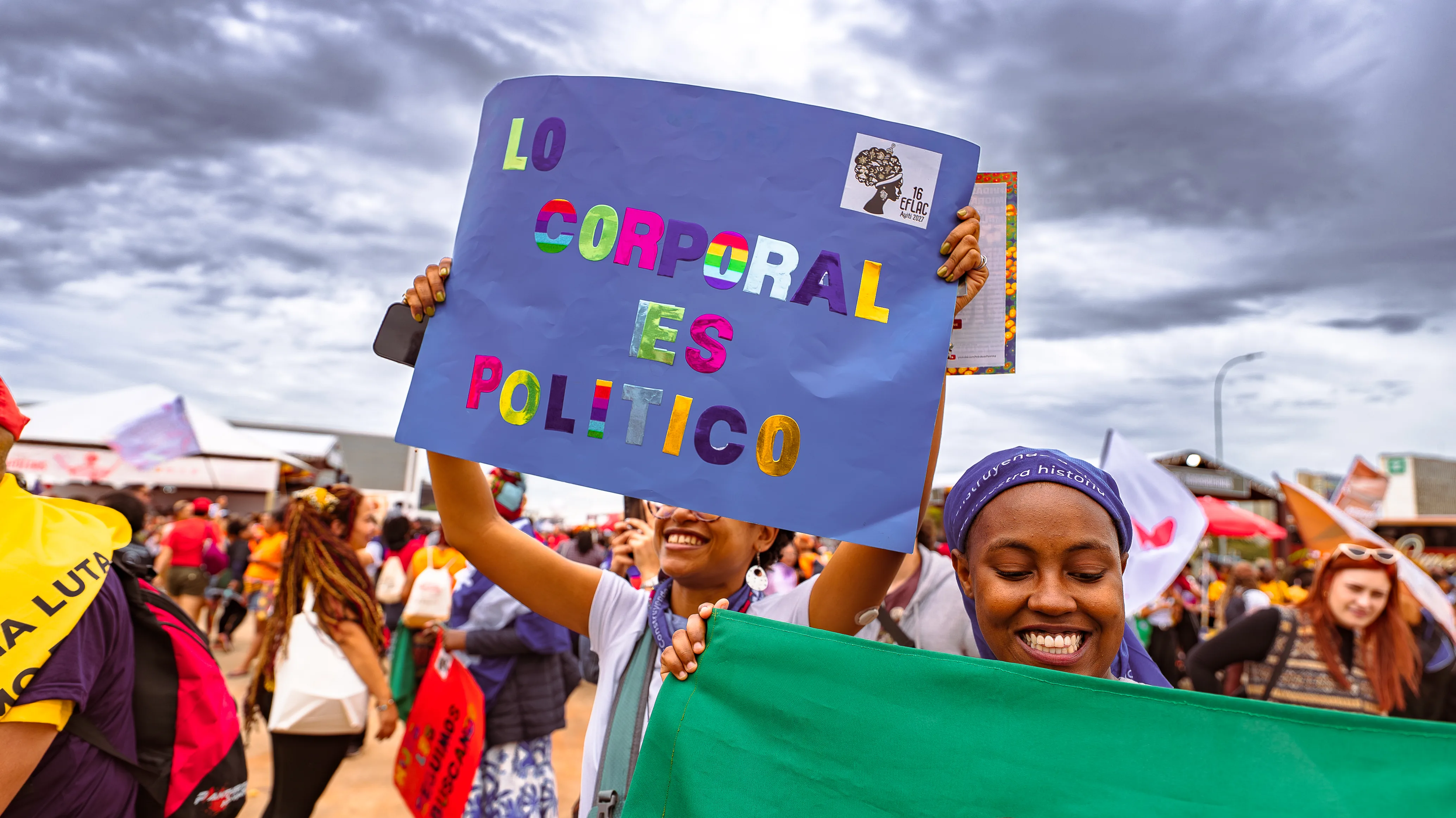 Mulher sorri enquanto segura cartaz azul com letras coloridas formando a frase “Lo corporal es político”. Ao fundo, outras pessoas caminham pela área do evento.