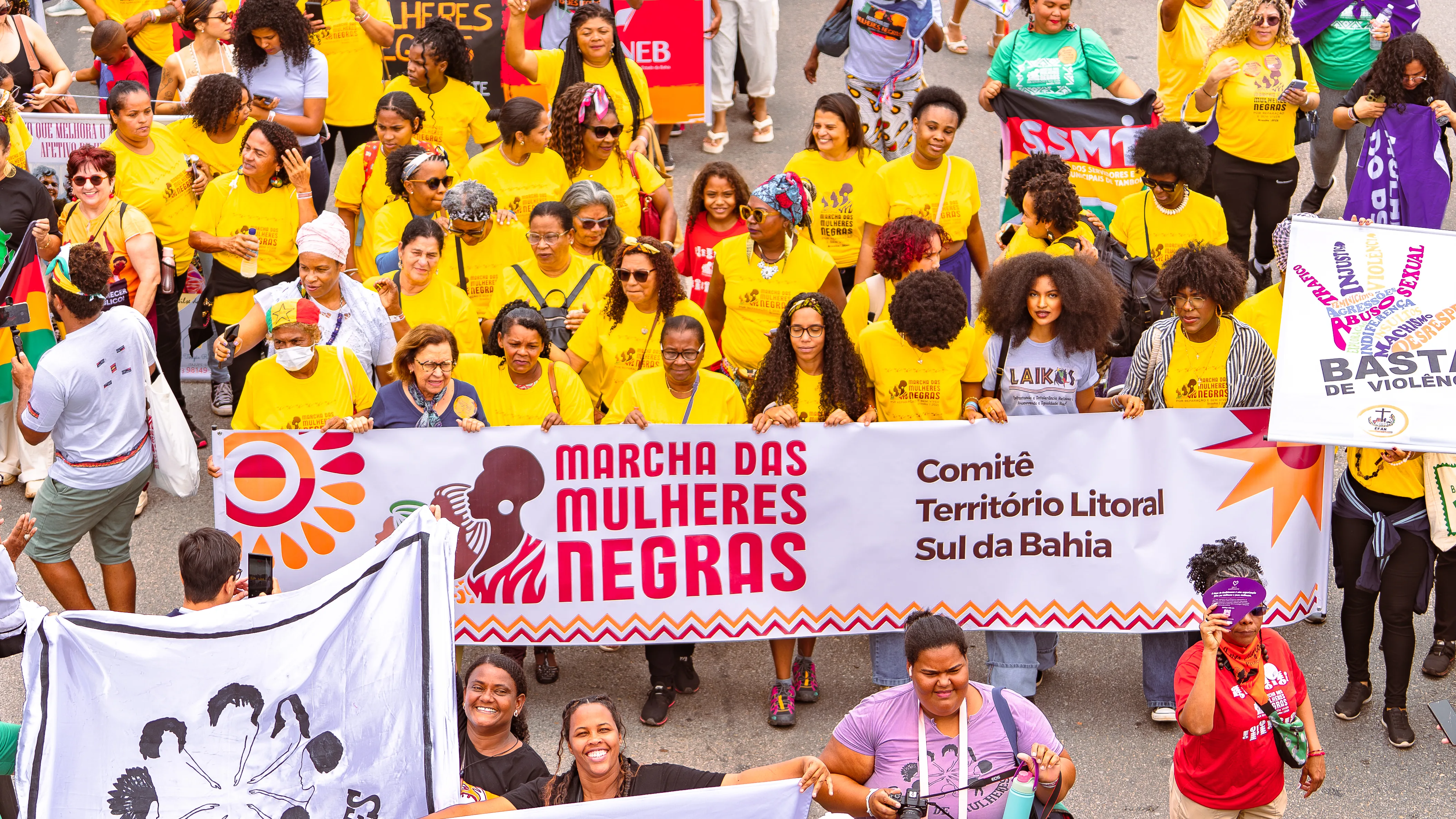 Grupo vestido majoritariamente com camisetas amarelas segura uma faixa branca com o nome da Marcha das Mulheres Negras e identificação do Comitê Território Litoral Sul da Bahia.