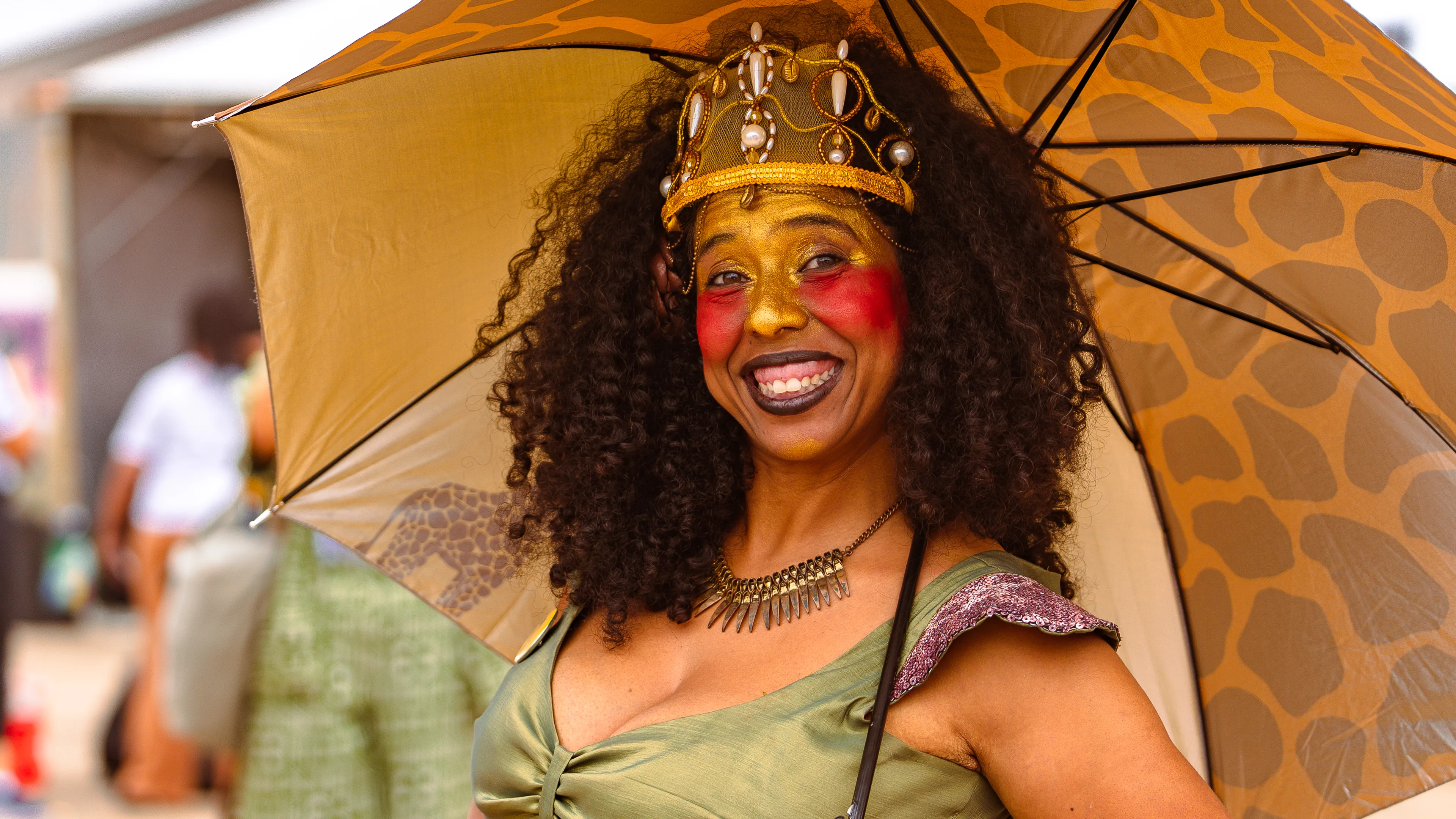 Mulher com maquiagem dourada no rosto, detalhe vermelho nas bochechas e coroa decorada com pérolas posa sorrindo sob um guarda-chuva marrom com estampa semelhante a pele de girafa. Ela usa vestido verde e colar metálico.