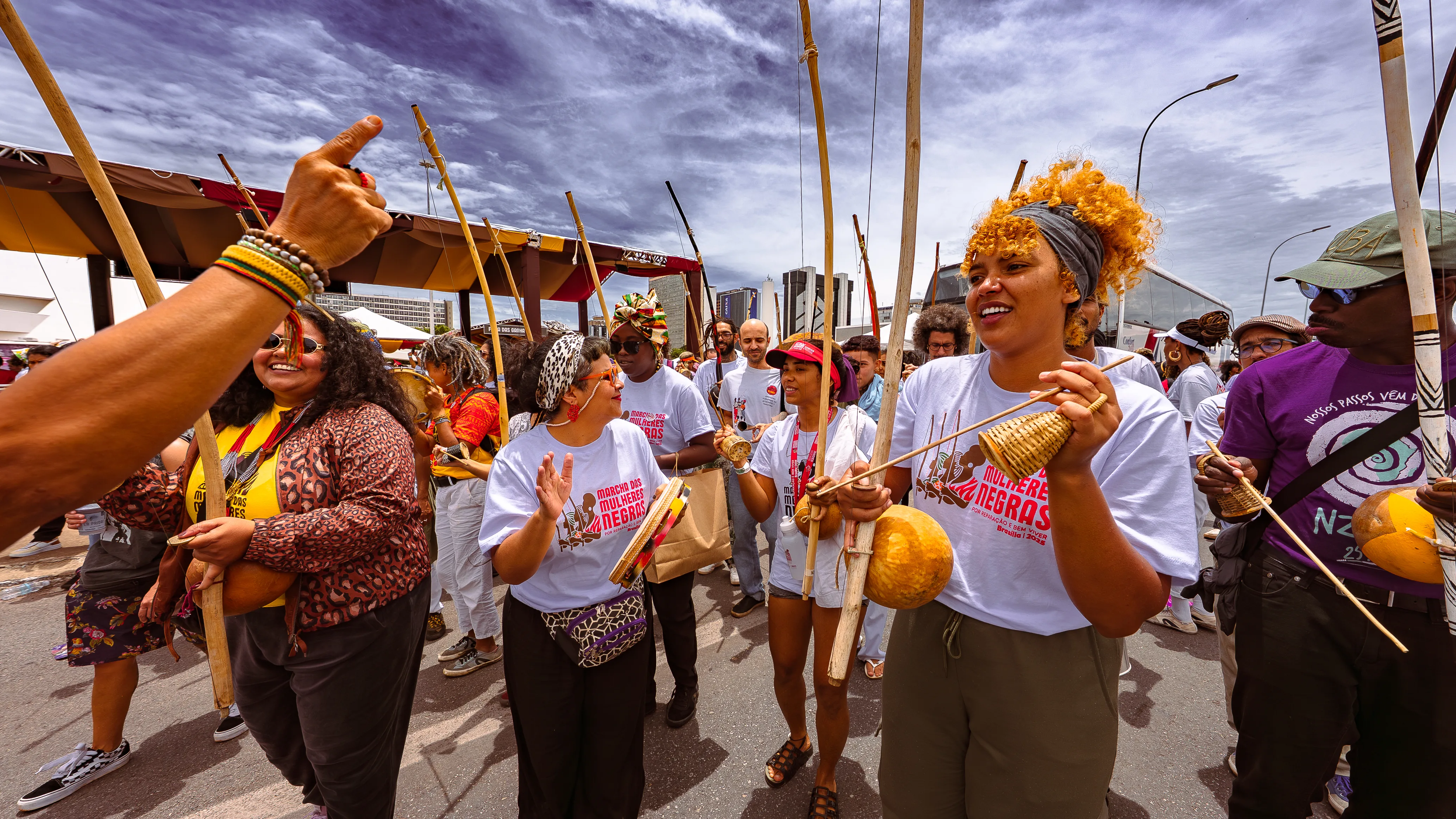 Um grupo de mulheres e homens segura berimbaus e instrumentos de percussão enquanto caminha na avenida, sorrindo e interagindo.