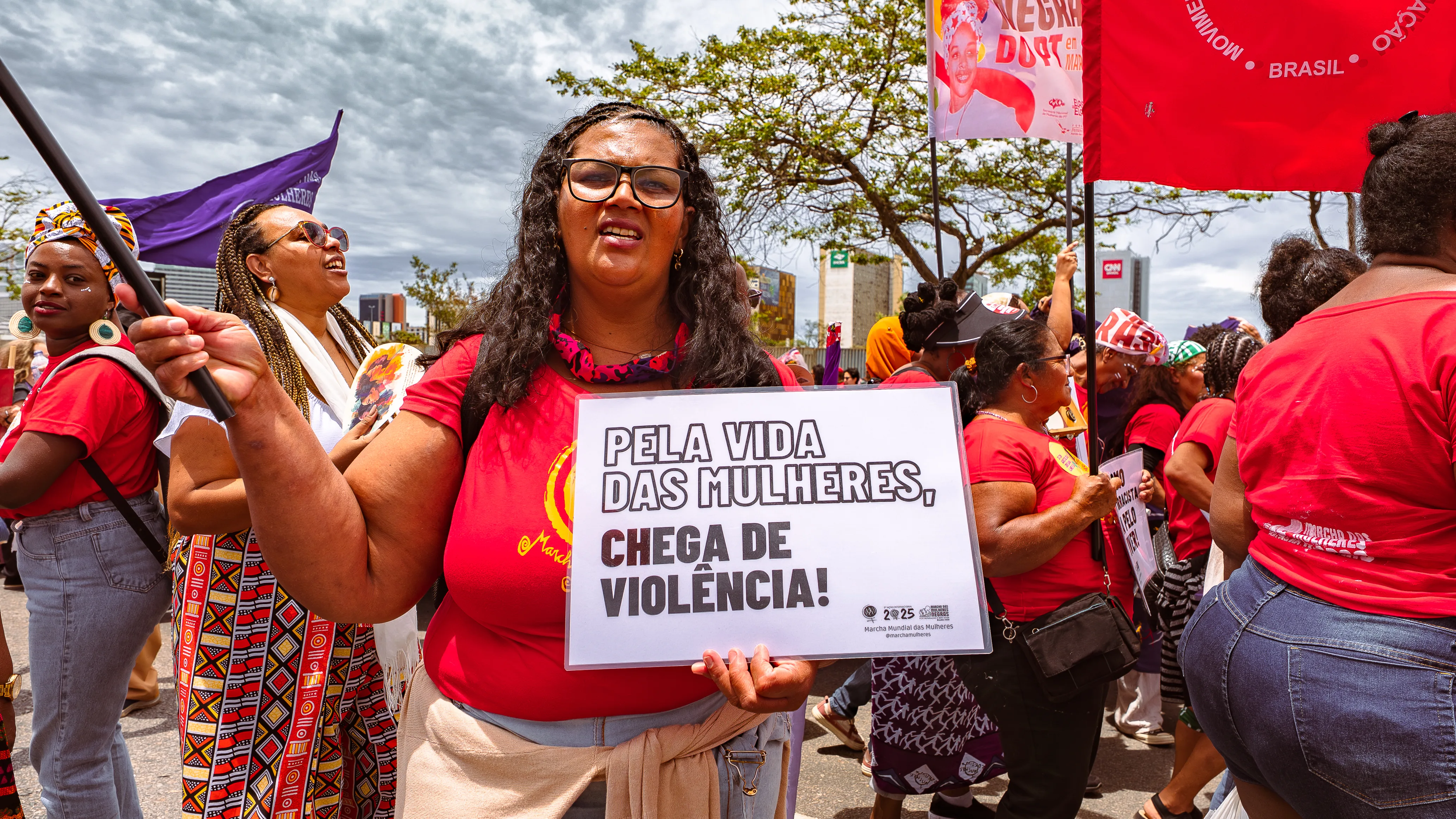 Mulher vestindo camiseta vermelha segura um cartaz com a frase “Pela vida das mulheres, chega de violência!”. Outras participantes caminham atrás dela.