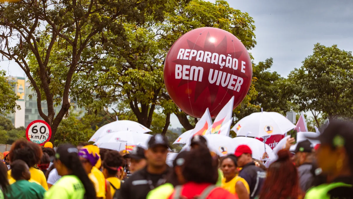 Um grande balão vermelho com a frase “Reparação e Bem Viver” paira acima da multidão, entre árvores e bandeiras. Pessoas caminham pela avenida durante o ato.