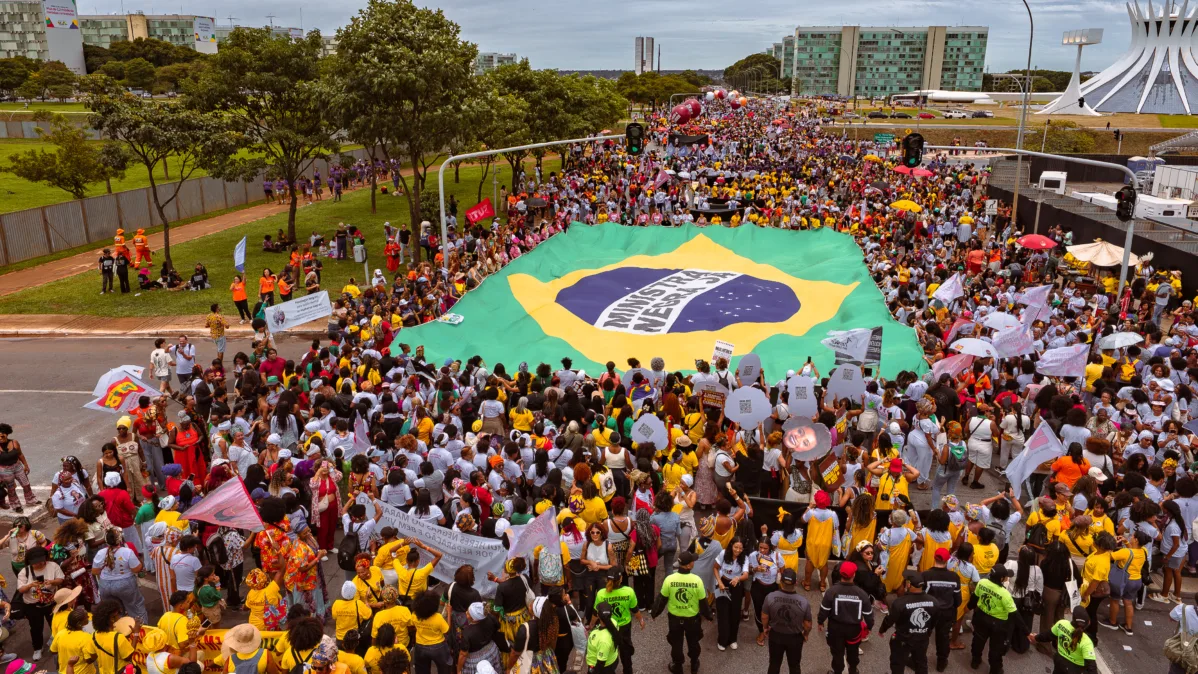 Uma grande multidão ocupa a avenida enquanto segura uma enorme bandeira com as cores do Brasil e a frase “Ministério da Igualdade Racial”. Prédios de Brasília aparecem ao fundo.