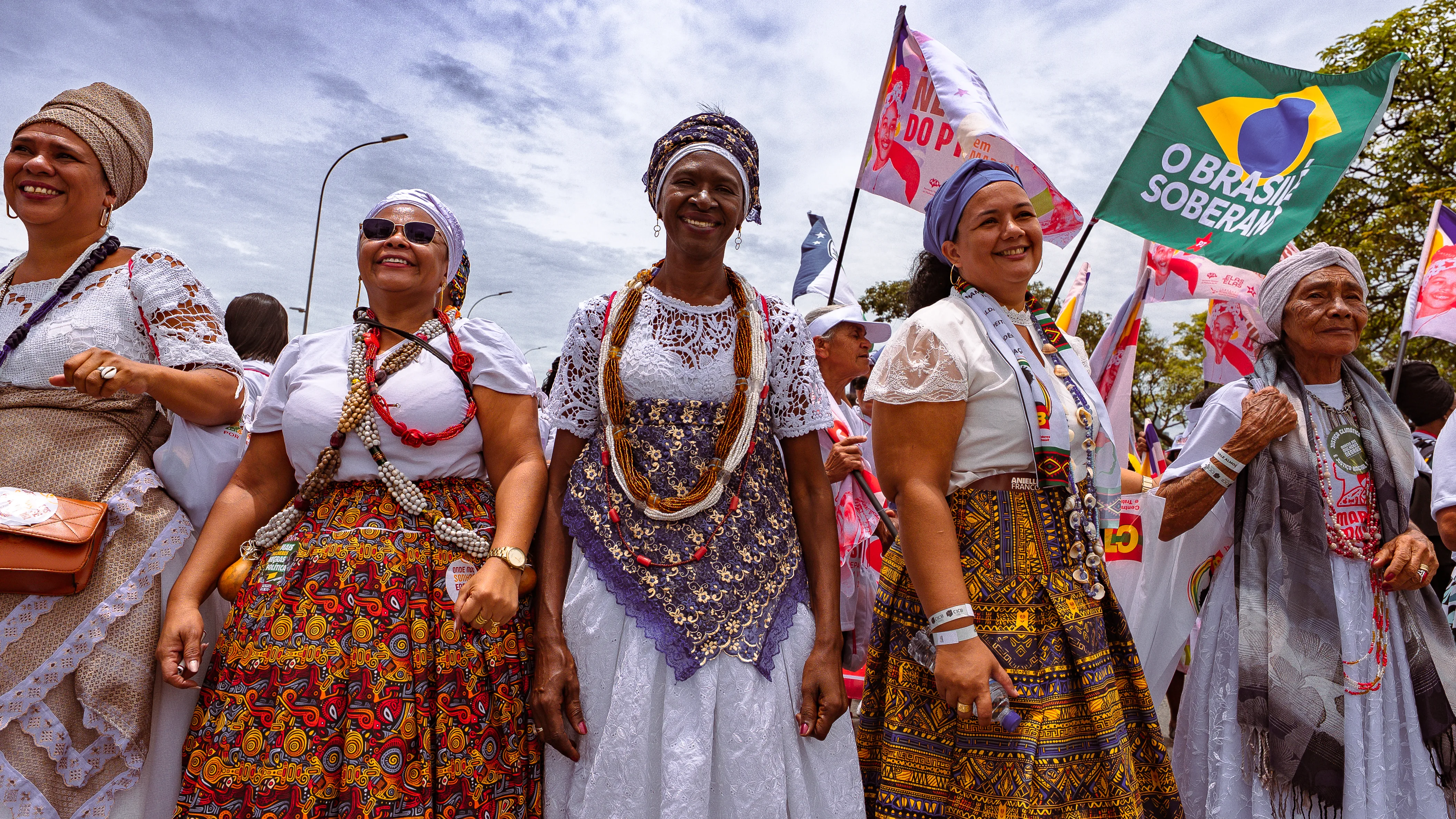 Mulheres vestidas com saias estampadas, rendas e turbantes caminham lado a lado, sorrindo. Bandeiras diversas aparecem ao fundo.