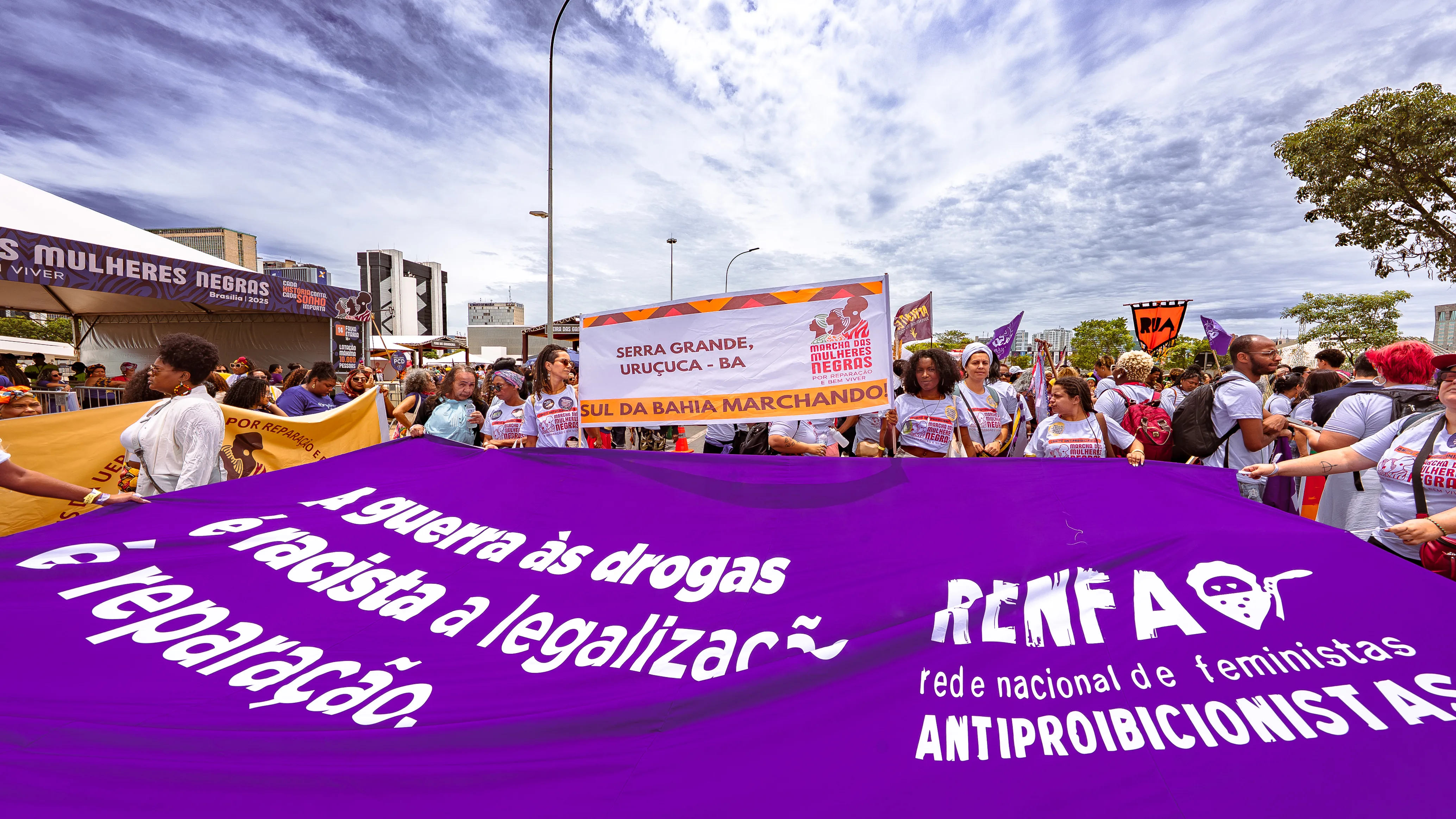 Manifestantes seguram uma grande faixa roxa da RENFA e outro banner da delegação de Serra Grande, Uruçuca–BA. O grupo ocupa a avenida com cartazes e camisetas da marcha.