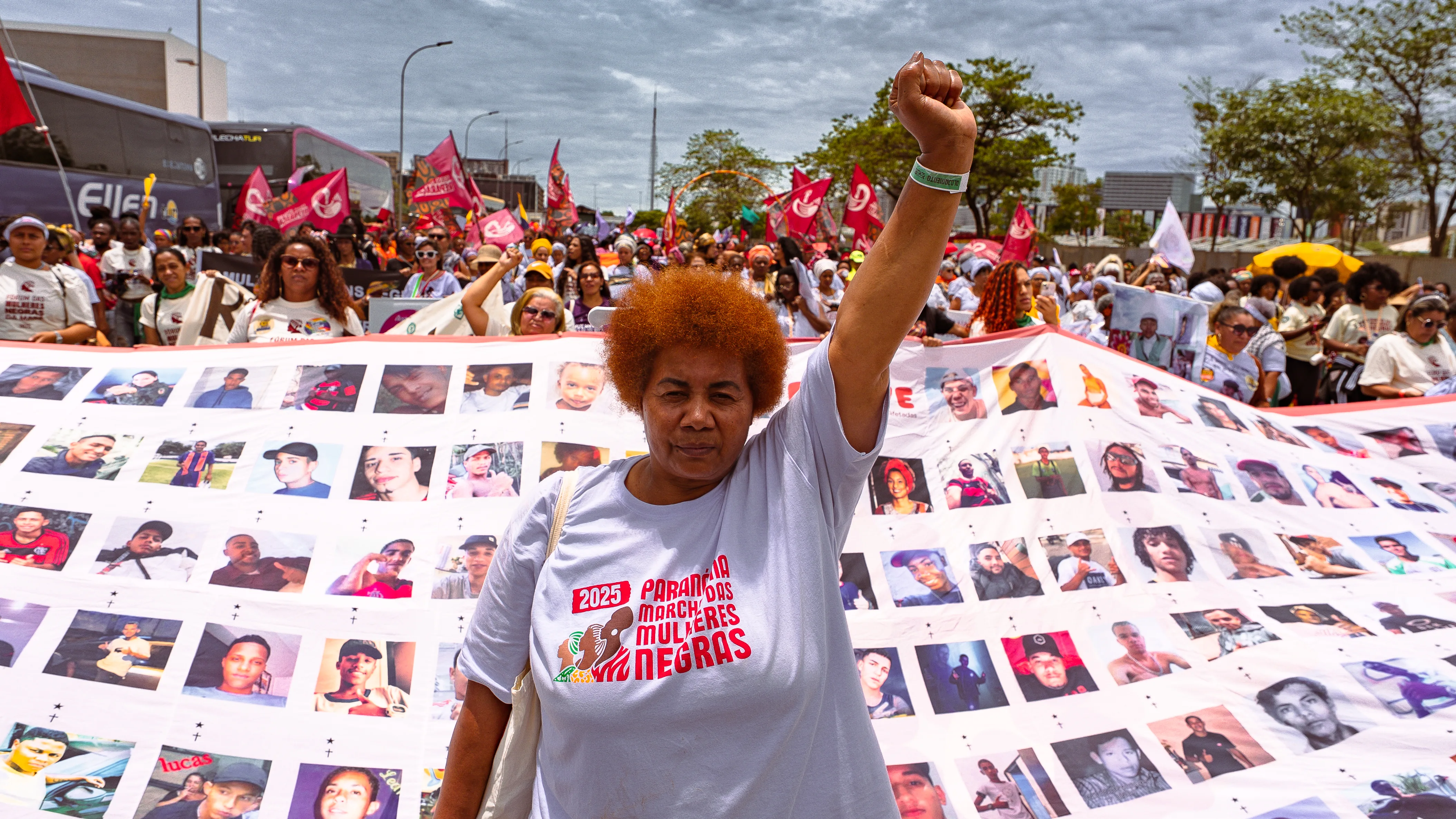 Uma mulher com camiseta da marcha ergue o punho enquanto está diante de um grande tecido repleto de fotos de homens e jovens. Atrás dela, uma multidão com bandeiras e cartazes preenche a avenida.