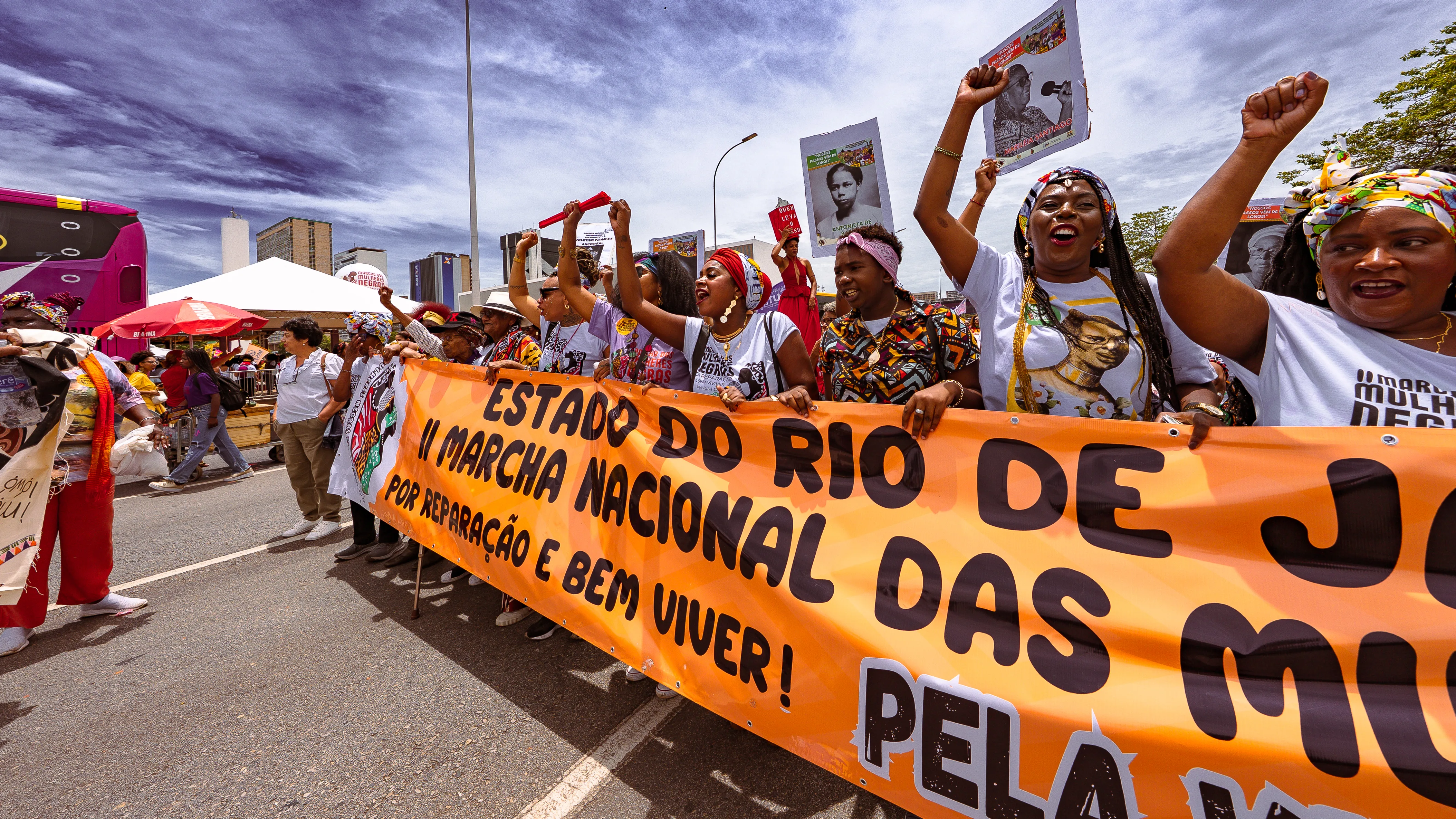 Um grupo de mulheres segura uma grande faixa laranja com inscrições sobre a Marcha Nacional das Mulheres Negras, levantando os braços e segurando cartazes. O céu está azul com nuvens e há prédios ao fundo.