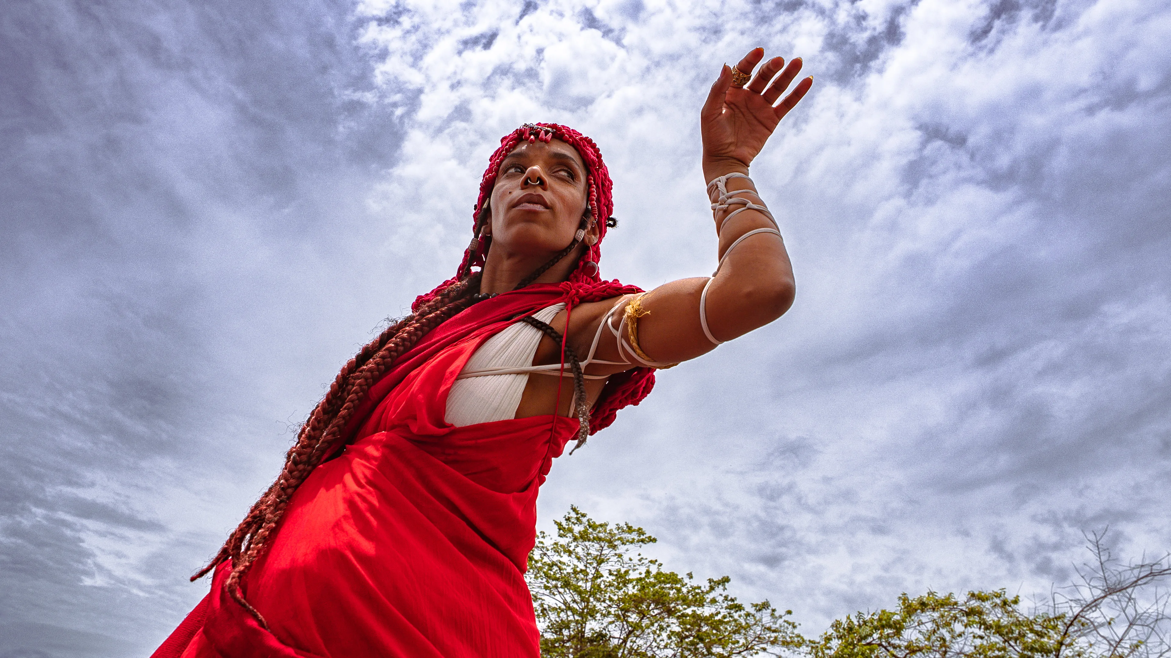 Mulher com longas tranças vermelhas e roupa vermelha ergue um braço durante uma apresentação. O céu nublado preenche o fundo.