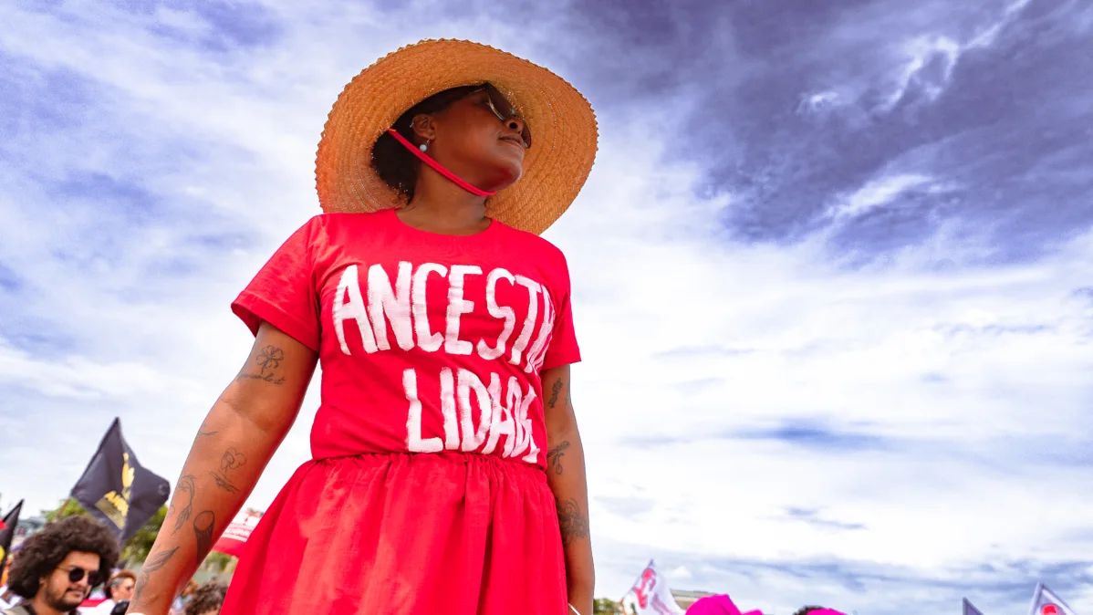 Mulher com chapéu de palha e camisa vermelha com a palavra “Ancestralidade” observa o ato. O céu aberto domina o fundo da imagem.