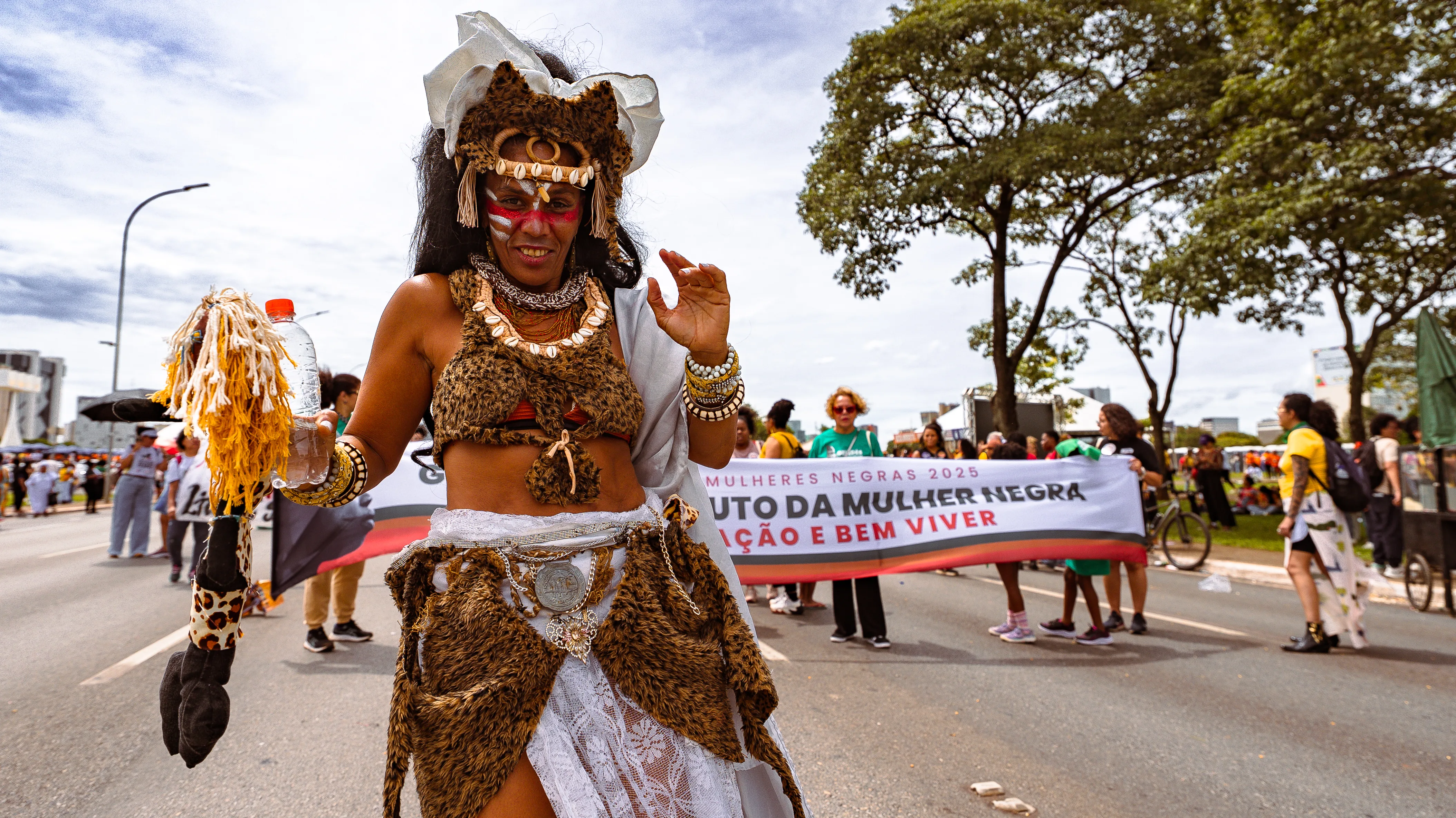 Uma mulher veste roupa com estampas que lembram pele de animal, adornada com colares e acessórios artesanais. Ao fundo, um grupo segura uma faixa da marcha.