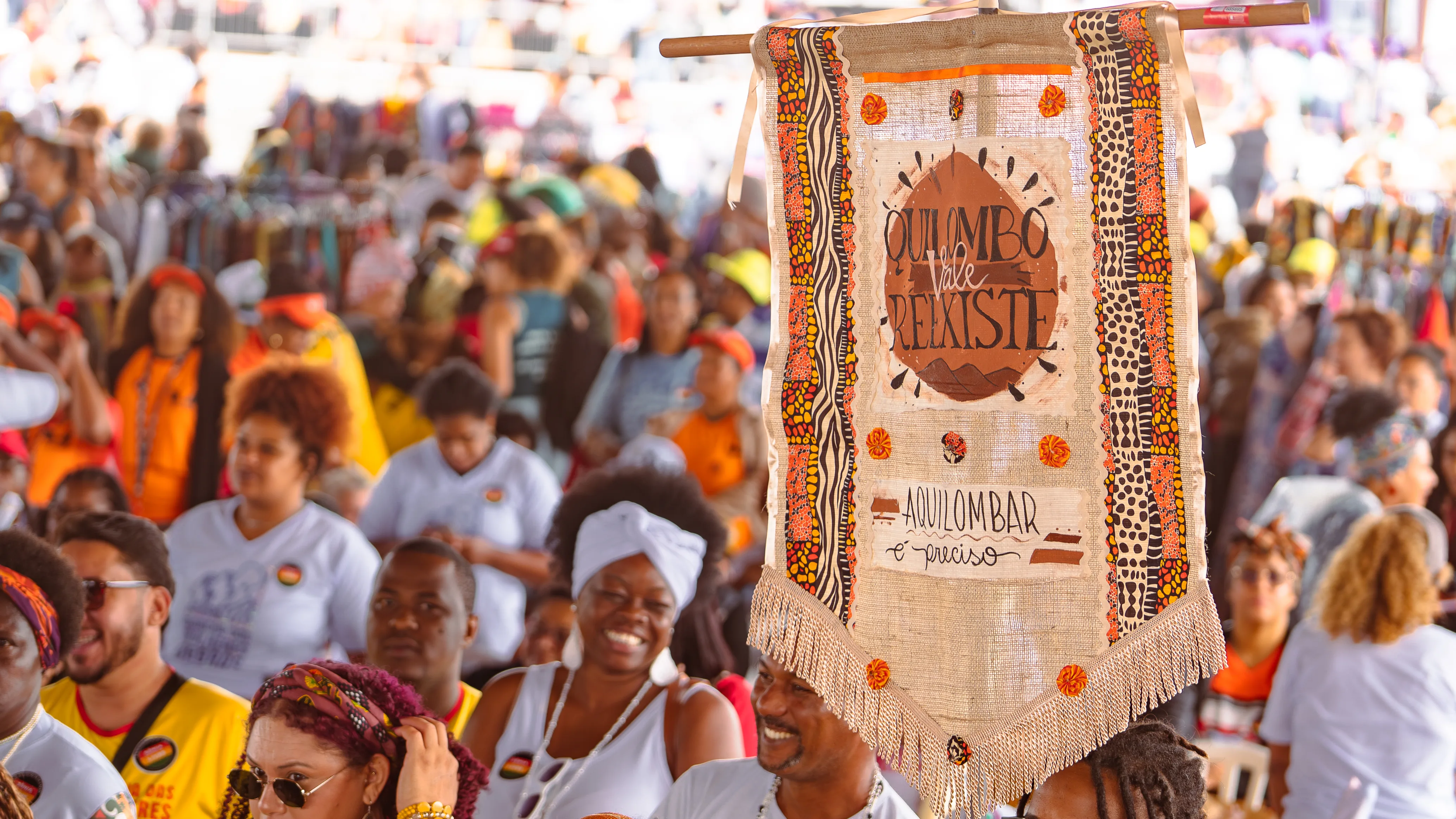 Estandarte de tecido com franjas e bordados coloridos exibe a frase “Quilombo Vale Resiste” acima de uma multidão movimentada.