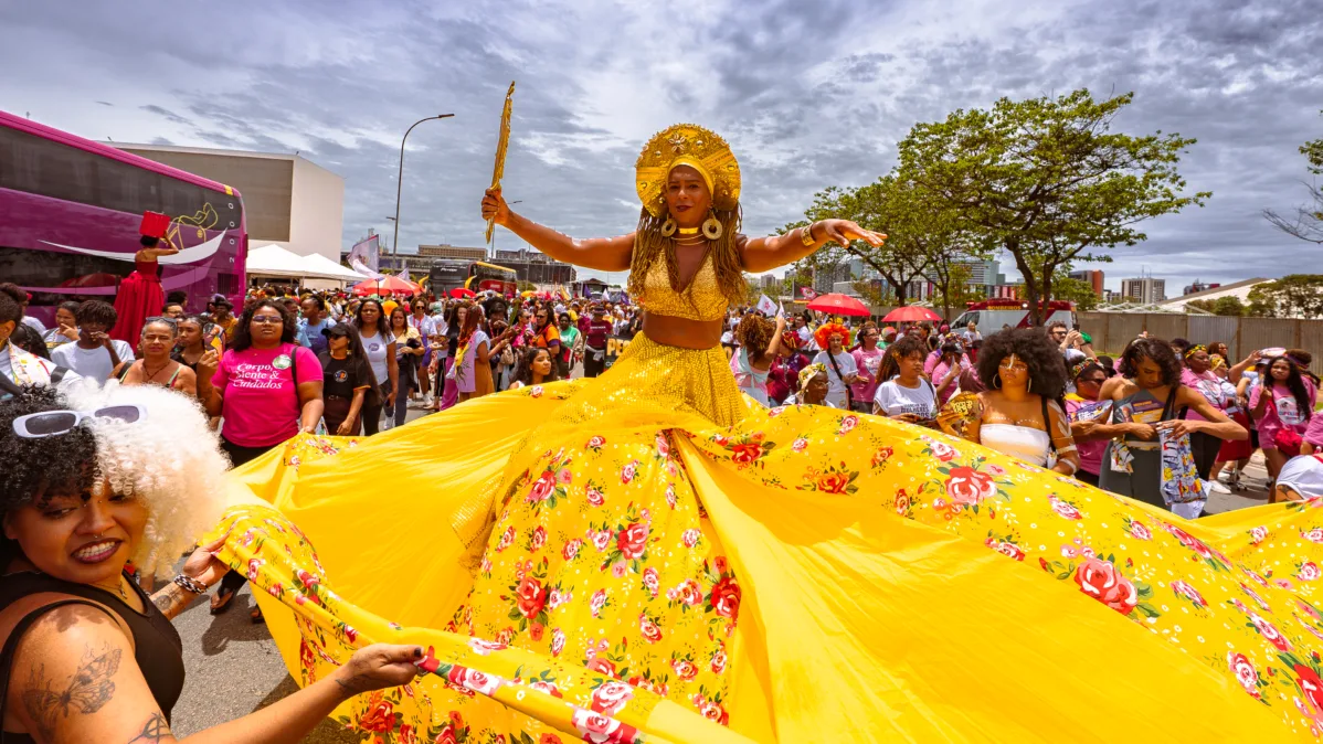 Uma mulher veste uma saia amarela muito ampla, estampada com flores coloridas, e um top brilhante. Ela ergue um objeto dourado enquanto outras mulheres ajudam a sustentar o tecido ao redor.
