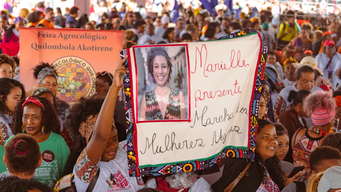 Pessoa ergue tecido com foto impressa de Marielle Franco e a frase “Marielle presente, Marcha das Mulheres Negras”. Ao fundo, há grande concentração de pessoas e um banner da feira quilombola.