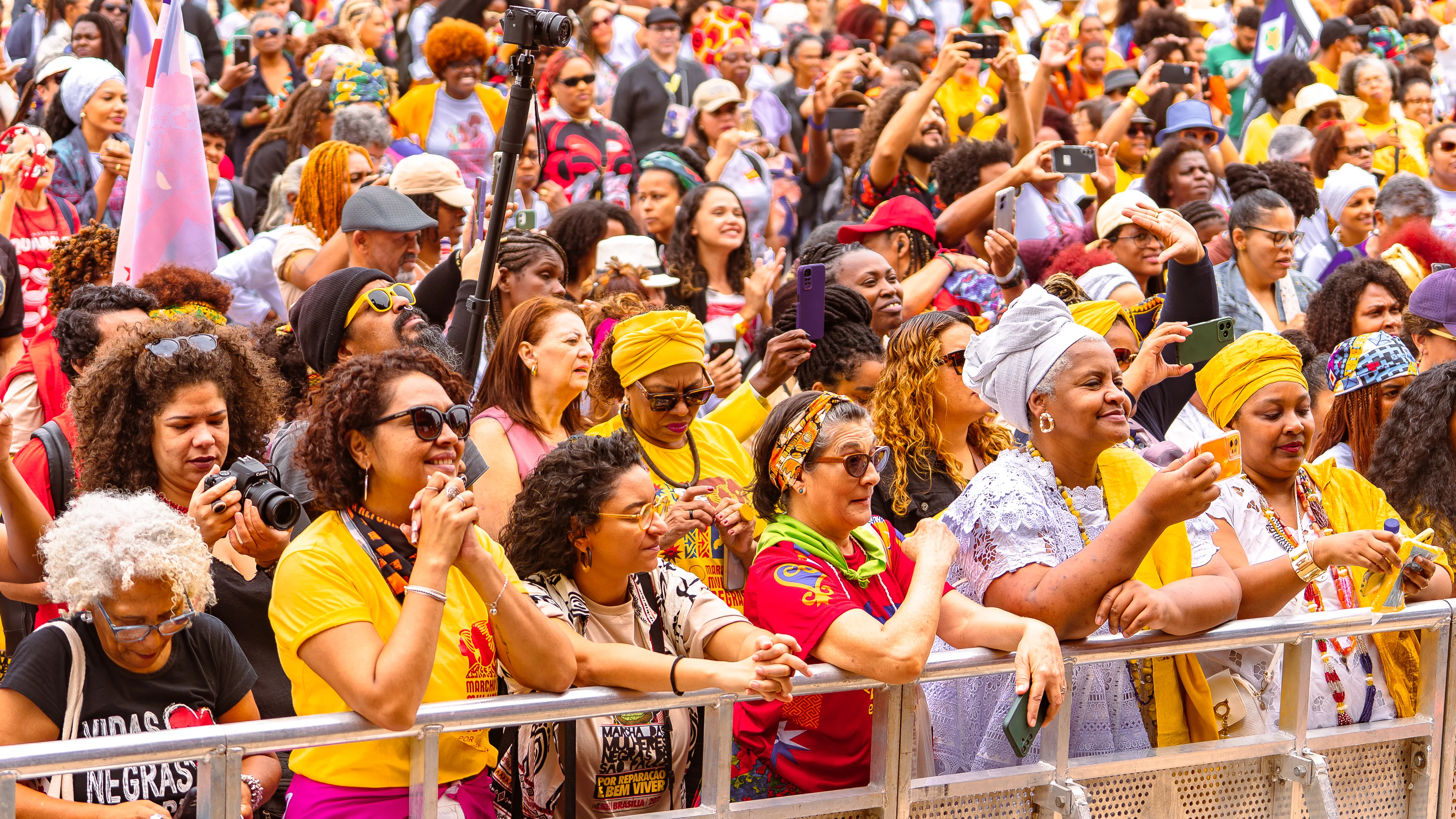 Diversas pessoas se aglomeram em frente a uma grade, algumas fotografando e outras observando atentamente. Há turbantes, camisetas amarelas e expressão geral de atenção.