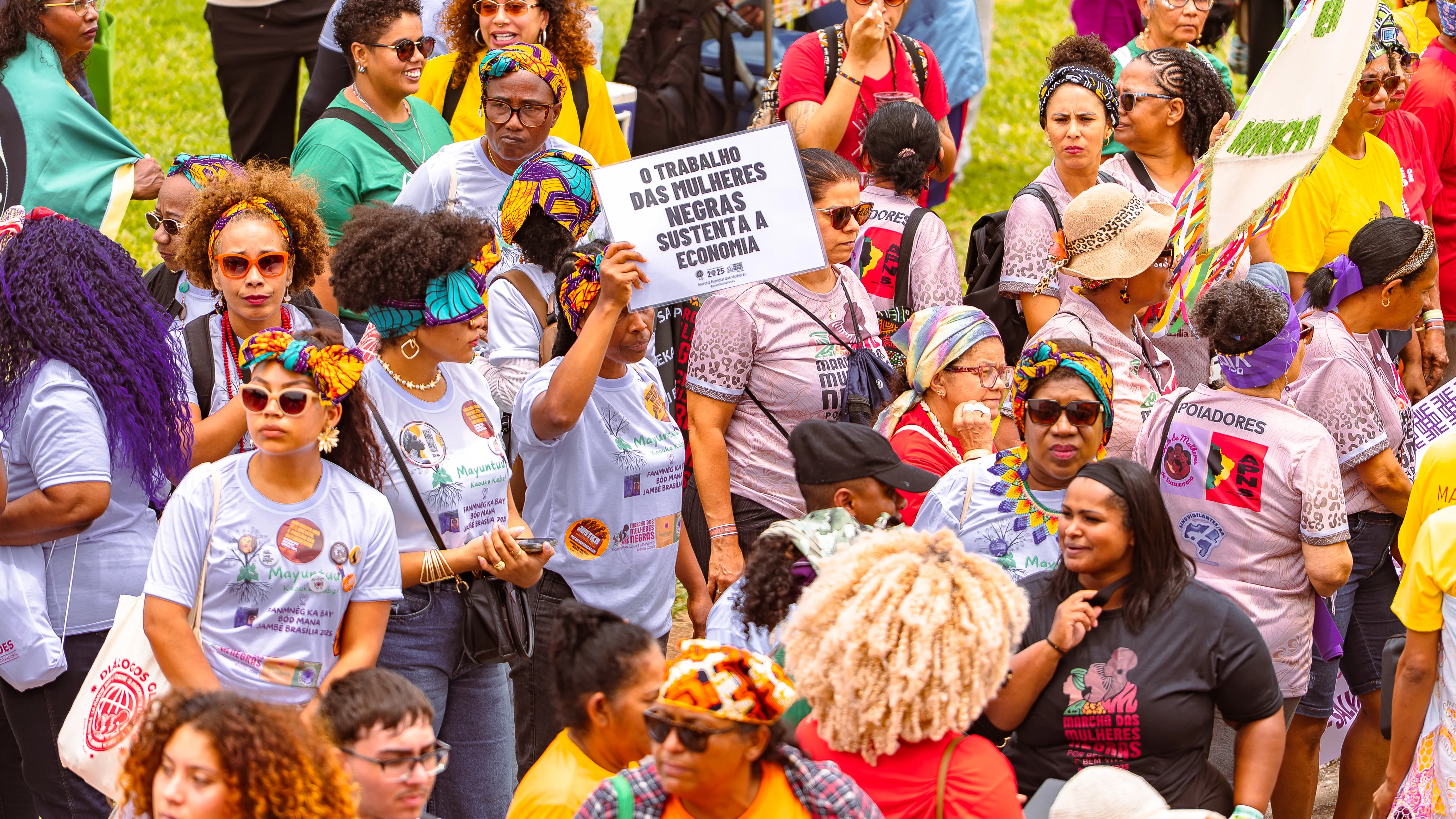 Um grupo de mulheres usa camisetas claras e turbantes coloridos; ao centro, uma delas ergue um cartaz branco com a frase “O trabalho das mulheres negras sustenta a economia”.