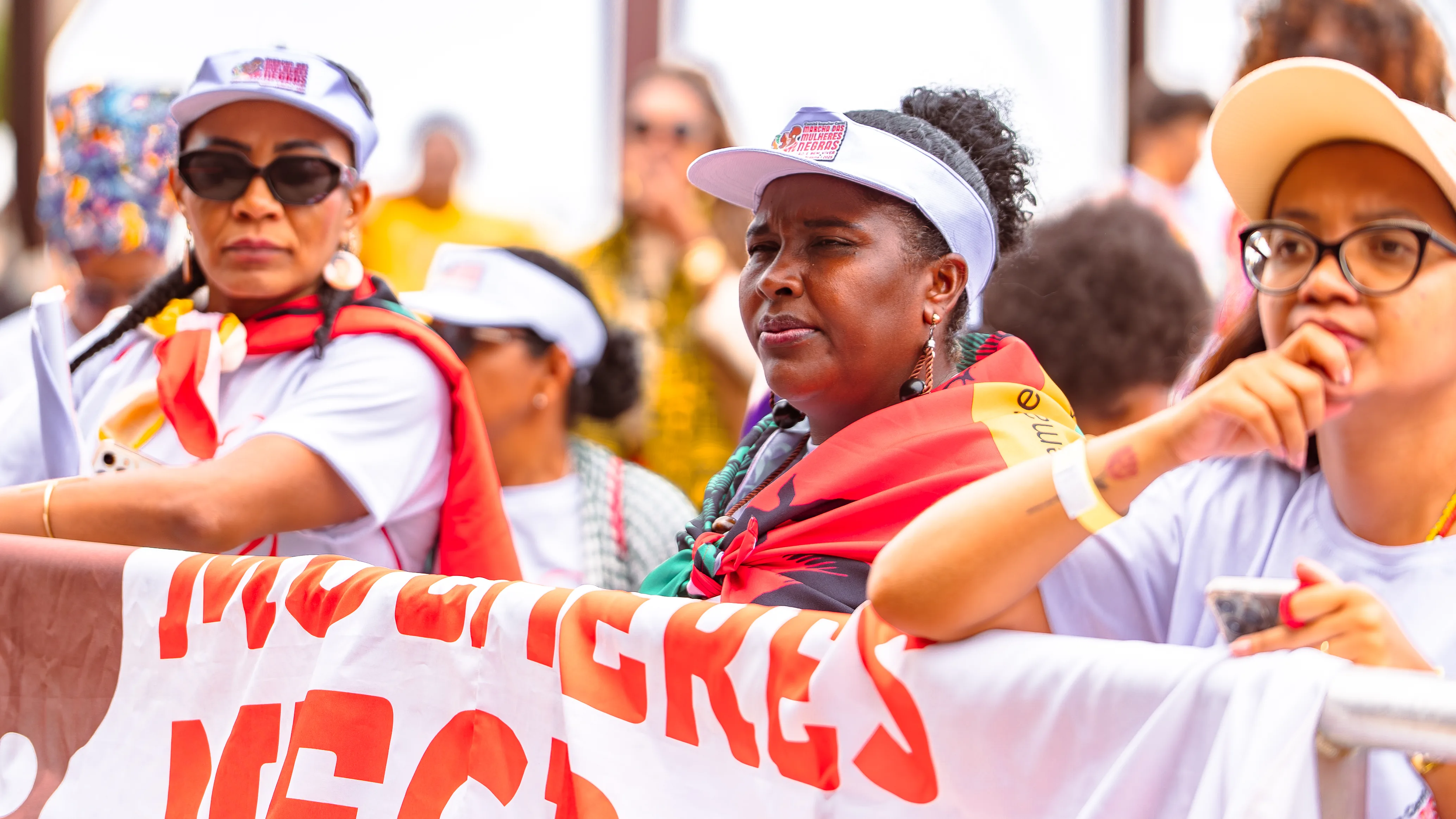 rês mulheres usam viseiras brancas e camisetas claras enquanto apoiam os braços sobre uma grande faixa branca e vermelha. Elas estão em ambiente externo, cercadas por outras pessoas ao fundo.