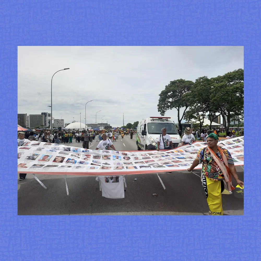 Mulheres caminham durante Marcha, segurando uma grande faixa repleta de fotos. Ao redor, há mais participantes, um carro e uma ambulância, além de prédios e árvores ao fundo sob céu nublado. A pessoa à frente à direita usa roupas coloridas e conduz uma das pontas da faixa.