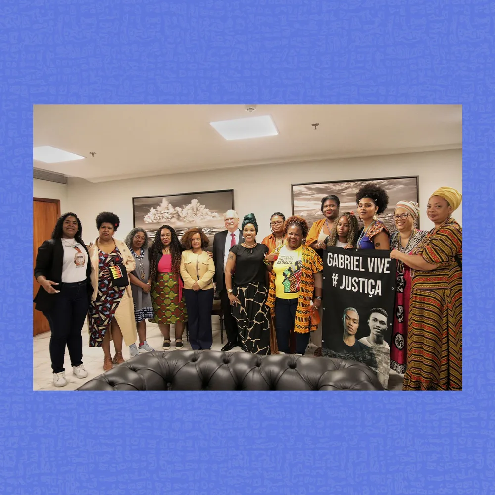 Grupo de cerca de quinze pessoas posando em uma sala iluminada, com duas fotos em preto e branco na parede ao fundo. A maioria veste roupas coloridas e algumas seguram uma faixa preta com o texto “Gabriel Vive". No primeiro plano, aparece a parte superior de um sofá de couro preto.