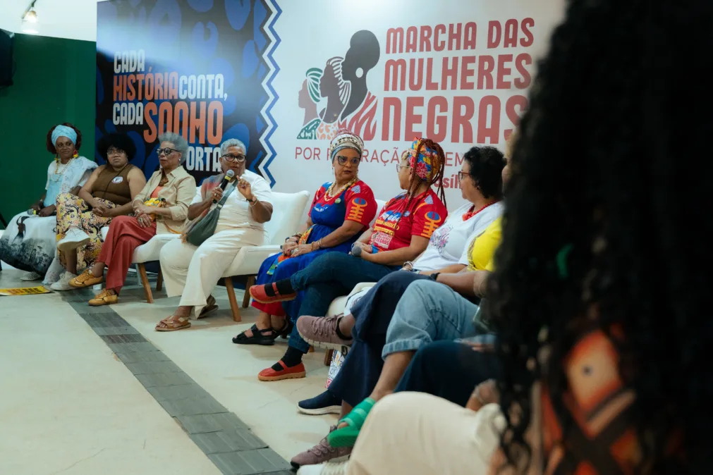 Mulheres negras sentadas, lado a lado. Uma das integrantes segura um microfone, enquanto as demais a observam. Ao fundo, um cartaz escrito "marcha das mulheres negras".