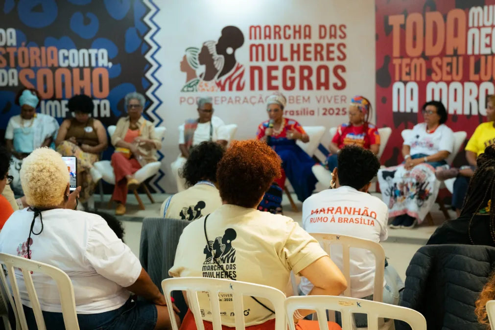 Em primeiro plano, mulheres sentadas em cadeiras olhando para o palco, onde mulheres negras estão sentadas, lado a lado. Ao fundo, um cartaz escrito "marcha das mulheres negras".