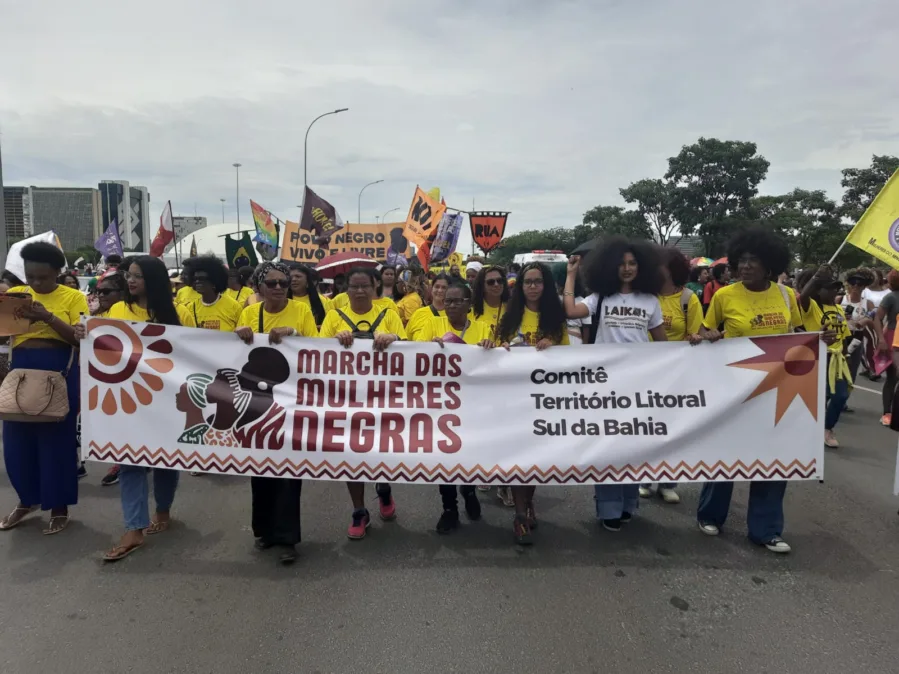 Grupo de mulheres negras marcha em avenida, muitas com camisetas amarelas. Na 1ª fileira, elas carregam uma grande faixa branca com o logotipo e o texto “Marcha das Mulheres Negras — Comitê Território Litoral Sul da Bahia”, ladeada por símbolos gráficos em marrom/laranja. Atrás, mais pessoas com cartazes e bandeiras coloridas (ex.: “Povo Negro Vivo e Livre”), árvores e prédios ao fundo sob céu nublado. Clima de mobilização, organização e orgulho.