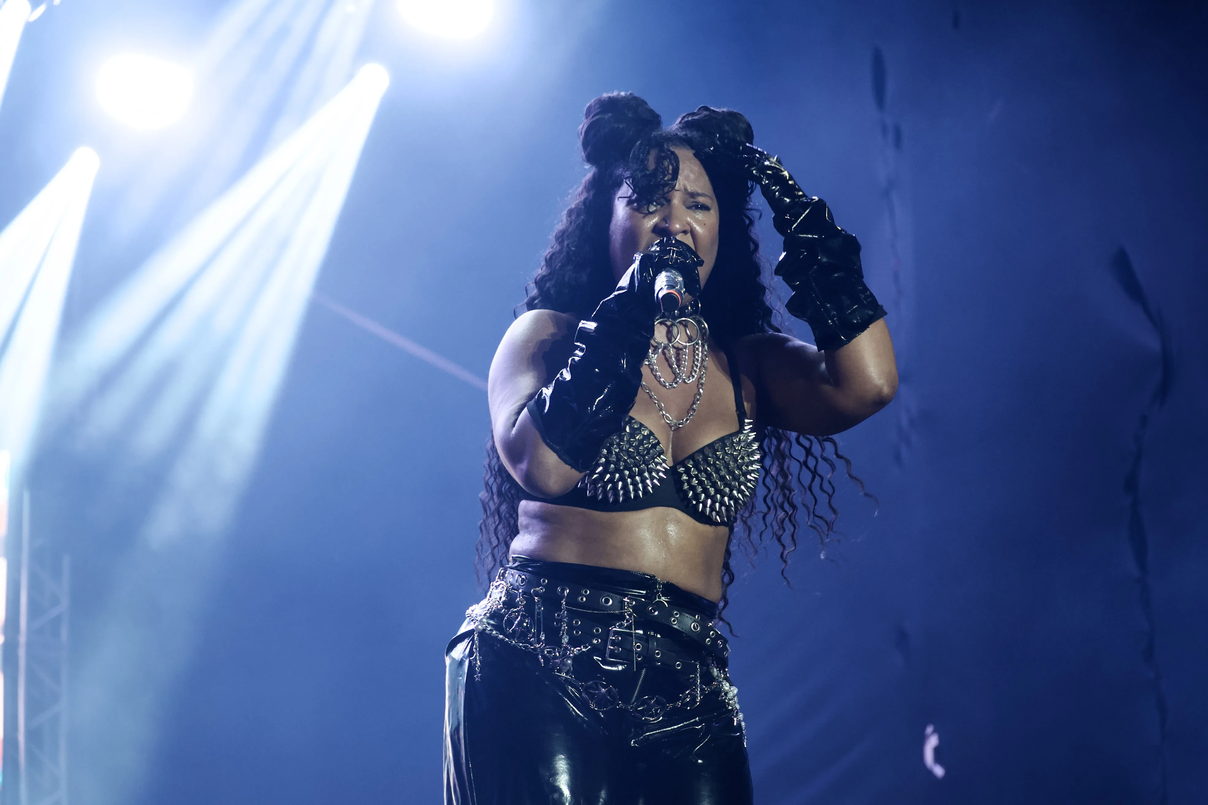 Mulher cantando no palco sob luzes fortes, usando um top preto com spikes metálicos, luvas pretas brilhantes e calça escura com cintos e correntes. Ela segura o microfone com uma das mãos enquanto a outra toca a cabeça. O fundo é escuro, iluminado por feixes de luz branca vindos de cima.