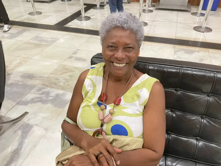Mulher negra idosa, de cabelo grisalho bem curto, sentada em um banco de couro preto. Sorri para a câmera, braços cruzados no colo, segurando um casaco bege. Veste vestido sem mangas branco com estampas verde-limão e um círculo azul; usa colar longo com contas grandes em tons de vermelho e rosa. Ambiente interno de saguão, com piso claro marmorizado e barreiras de fila ao fundo.