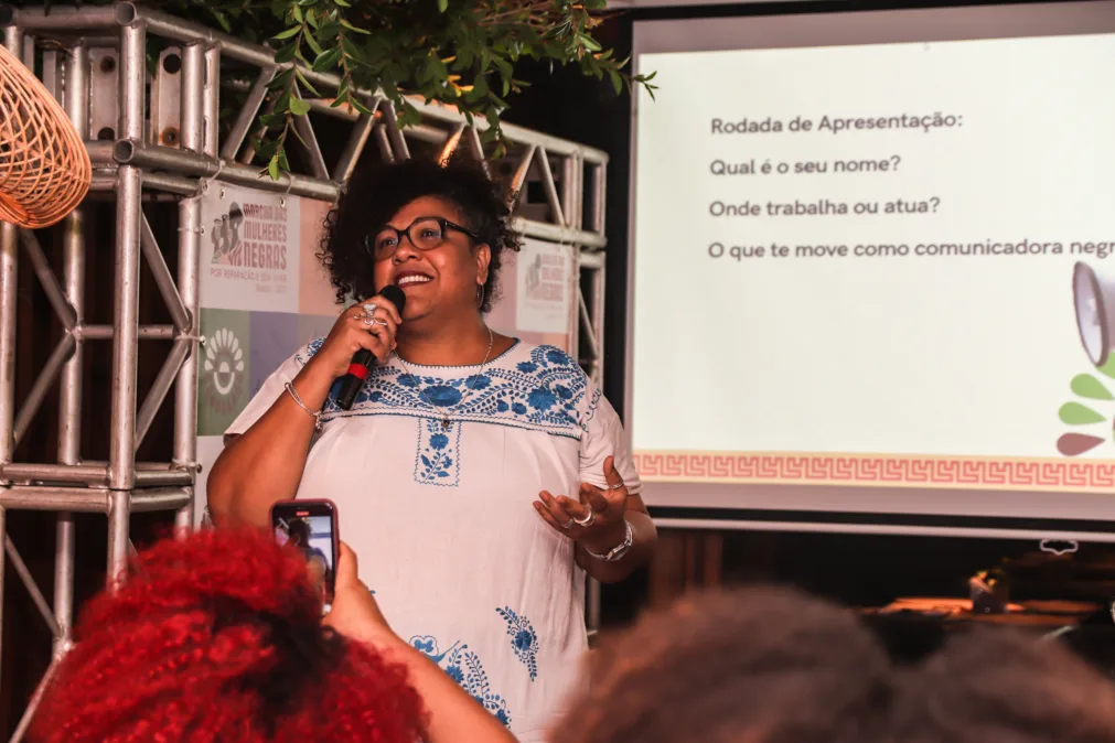 Mulher negra com cabelos cacheados e óculos fala ao microfone diante de um telão. Veste bata branca com bordados azuis e usa anéis, bracelete e colar. À esquerda, estrutura de truss com um banner da Marcha das Mulheres Negras. No telão, lê-se: “Rodada de Apresentação: Qual é o seu nome? Onde trabalha ou atua? O que te move como comunicadora negra?”. Na frente, público com celular erguito registra a fala. Ambiente interno, iluminação quente, clima de encontro/formação.