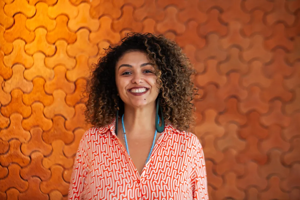 Retrato de uma mulher sorrindo, em pé, de frente. Cabelo cacheado médio, volumoso, com mechas claras. Usa camisa longa vermelha com estampa geométrica branca, mangas dobradas e mãos nos bolsos laterais. Brinco verde em argola alongada e colar azul fino. Ao fundo, parede laranja com relevo em formas curvas, iluminação quente. Expressão acolhedora e confiante.