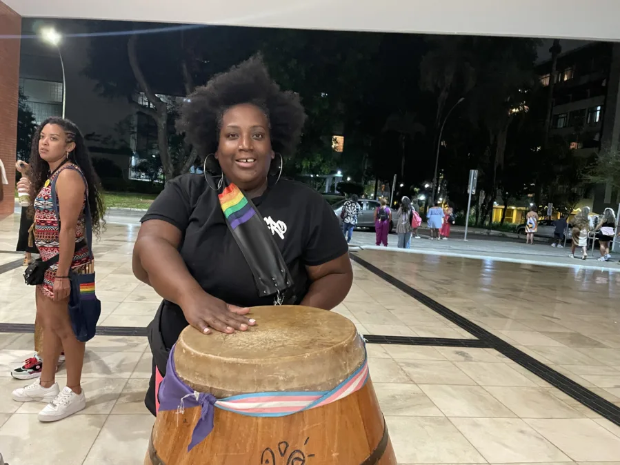 Mulher negra com cabelo Black, sorrindo, posa com a mão apoiada sobre um tambor de madeira grande. Ela usa camiseta preta, argolas prateadas e bolsa transversal com faixa/patch de arco-íris. No tambor há fitas nas cores da bandeira trans e uma faixa roxa. Cena noturna em área aberta com piso claro; ao fundo, pessoas conversam e caminham, árvores e prédios iluminados.