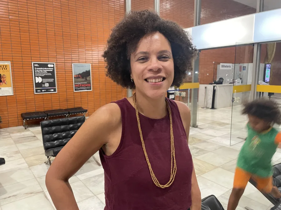 Mulher negra de cabelo crespo curto sorri para a câmera, de pé, mãos na cintura. Usa blusa sem mangas vinho e colar longo de miçangas douradas. Ambiente interno com paredes de tijolinhos laranja, bancos pretos e pôsteres ao fundo; portas de vidro com letreiro “Bilheteria”. À direita, uma criança passa correndo, levemente borrada.