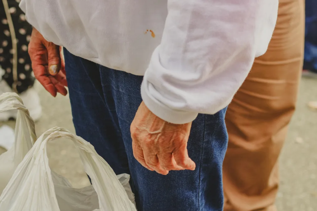 Mãos de pessoa idosa com moletom branco e calça azul; segura sacolas plásticas. Close em fila, rua ao fundo.