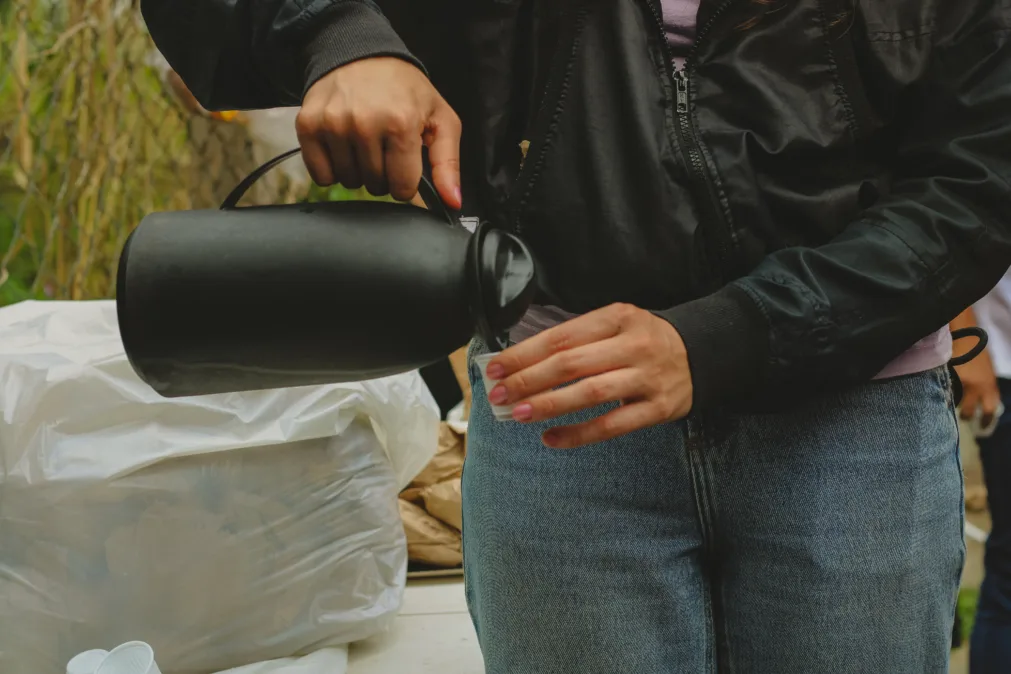 Pessoa com jaqueta preta e calça jeans derramando café de uma garrafinha preta em um copo descartável, com sacos plásticos ao fundo.