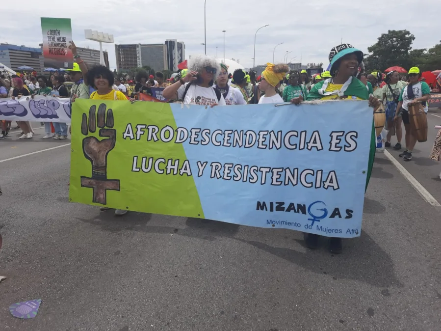 Mulheres marcham em avenida larga segurando um grande banner bicolor (verde e azul-claro) com o texto: “AFRODESCENDENCIA ES LUCHA Y RESISTENCIA”. À esquerda do banner, há um símbolo de punho erguido combinado ao símbolo feminino. No canto inferior direito, lê-se “MIZANGAS – Movimiento de Mujeres Afro”. As participantes usam roupas e turbantes coloridos; ao fundo, mais pessoas com faixas, tambores e bandeiras. Céu nublado e prédios ao fundo.