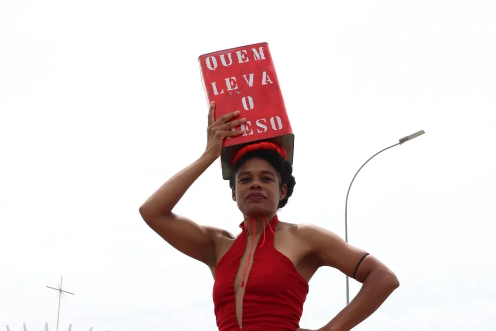 Mulher negra com uma lata na cabeça escrito "quem leva o peso". Ela veste vermelho. 