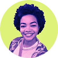 Mulher negra de cabelos crespos e curtos. Ela está sorrindo e usando uma blusa branca com sobreposição estampada.
