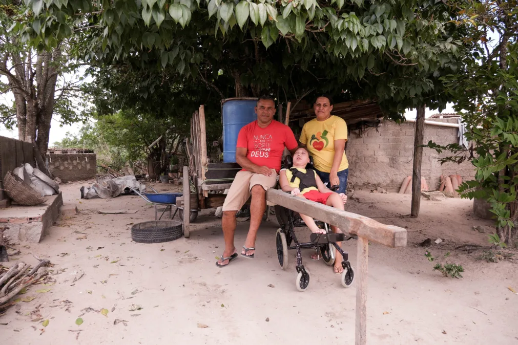 De dia, em quintal de terra sob copa de árvore, um homem de camisa vermelha senta na carroça; ao lado, uma mulher de camiseta amarela. À frente, uma criança em cadeira de rodas, com apoio de cabeça, olha para cima. Atrás deles há um tambor azul sobre a carroça e, ao redor, ferramentas, pneus e um muro