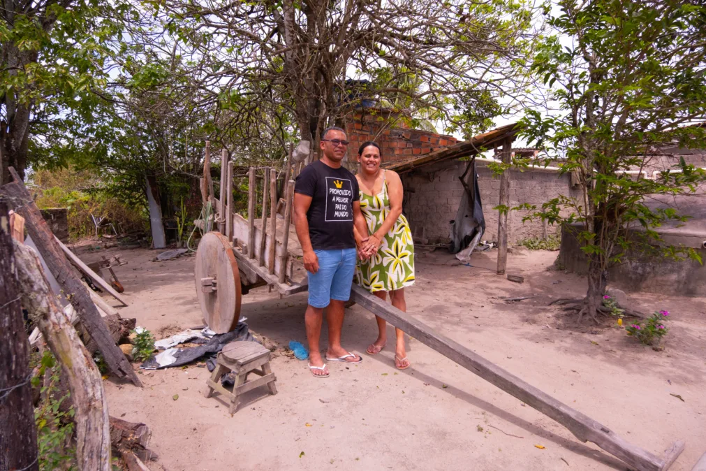 Em pátio de terra, um casal posa de mãos dadas ao lado de uma carroça de madeira com rodas grossas e vara longa voltada para a câmera. O homem usa camiseta preta e bermuda jeans; a mulher, vestido verde com estampa floral. Ao fundo, árvores ralas, um galpão simples e paredes de alvenaria.
