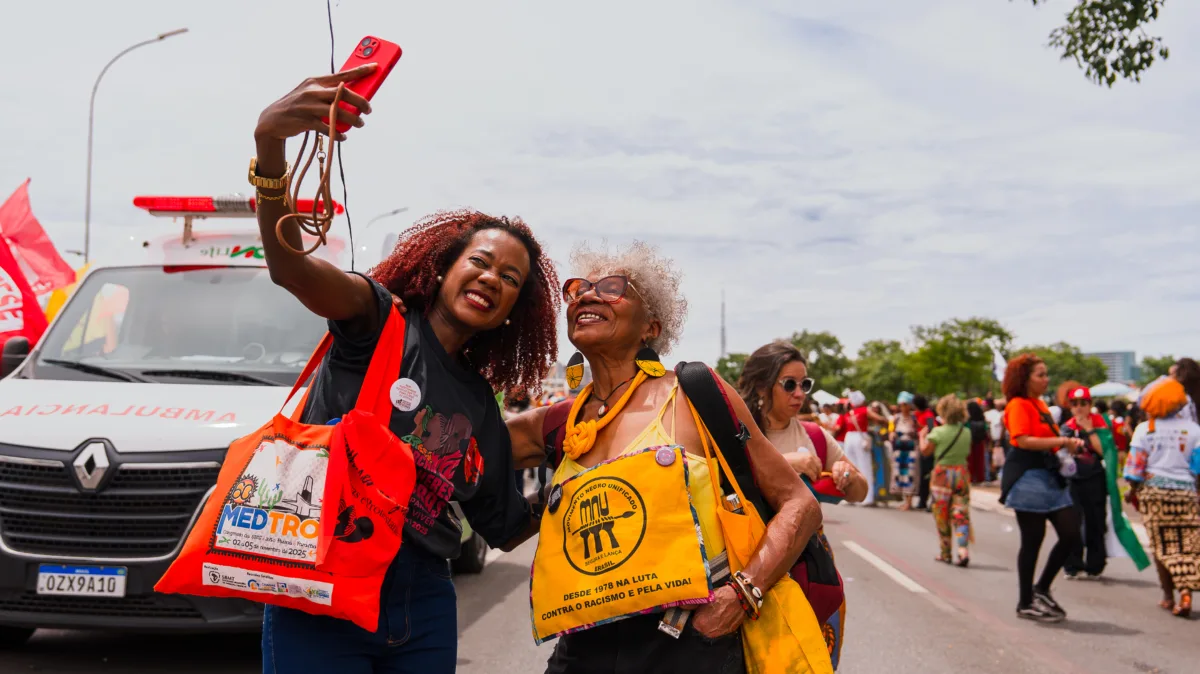 Duas mulheres negras, uma com camiseta preta e outra com camiseta amarela do MNU, tiram selfie sorrindo durante a marcha. Elas carregam bolsas de tecido com estampas do evento; atrás, ambulância e um fluxo de manifestantes na avenida. Atmosfera festiva e combativa. Foto: Marina Gadelha.