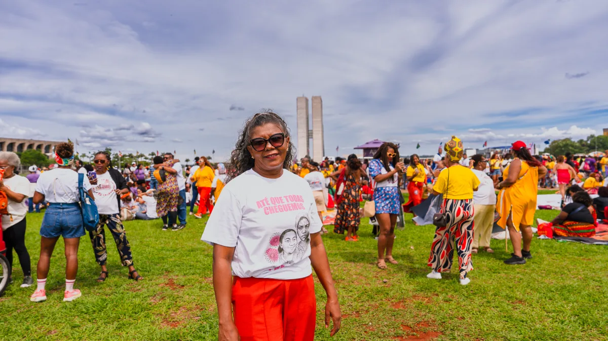 Mulher negra de cabelos grisalhos e óculos escuros posando de frente para a câmera. Veste a camiseta branca com ilustrações em rosa e a frase “ATÉ QUE TODAS CHEGUEMOS”. O Congresso Nacional surge desfocado atrás dela, com pessoas circulando no gramado.